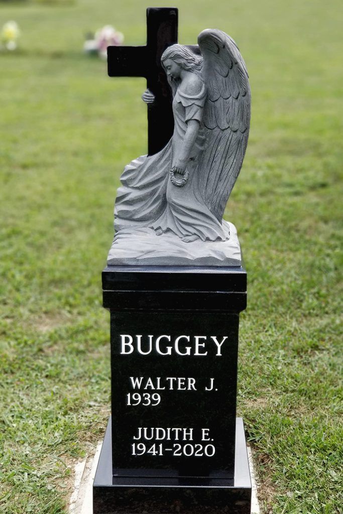 Gray angel statue atop black memorial gravestone in a grassy cemetery, with engraved names and dates.