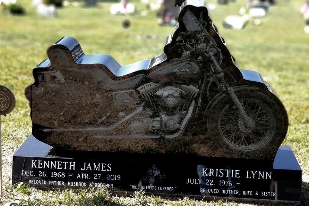 Motorcycle-shaped gravestone in a cemetery, black base with engraved names and dates.