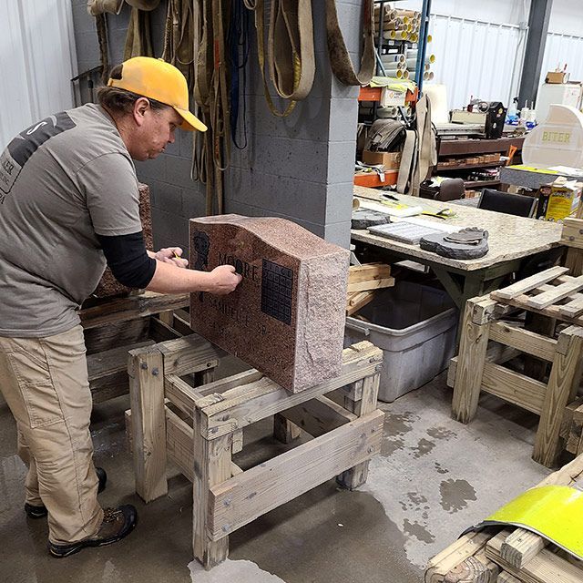 Worker in a yellow cap shaping a large stone block in a workshop with tools and equipment nearby