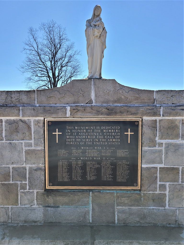 Statue of a saint atop a stone wall above a memorial plaque with crosses, outdoors under a blue sky