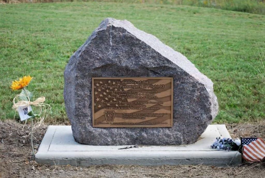 Memorial stone with bronze plaque on a concrete base in a grassy cemetery, with flowers and an American flag.