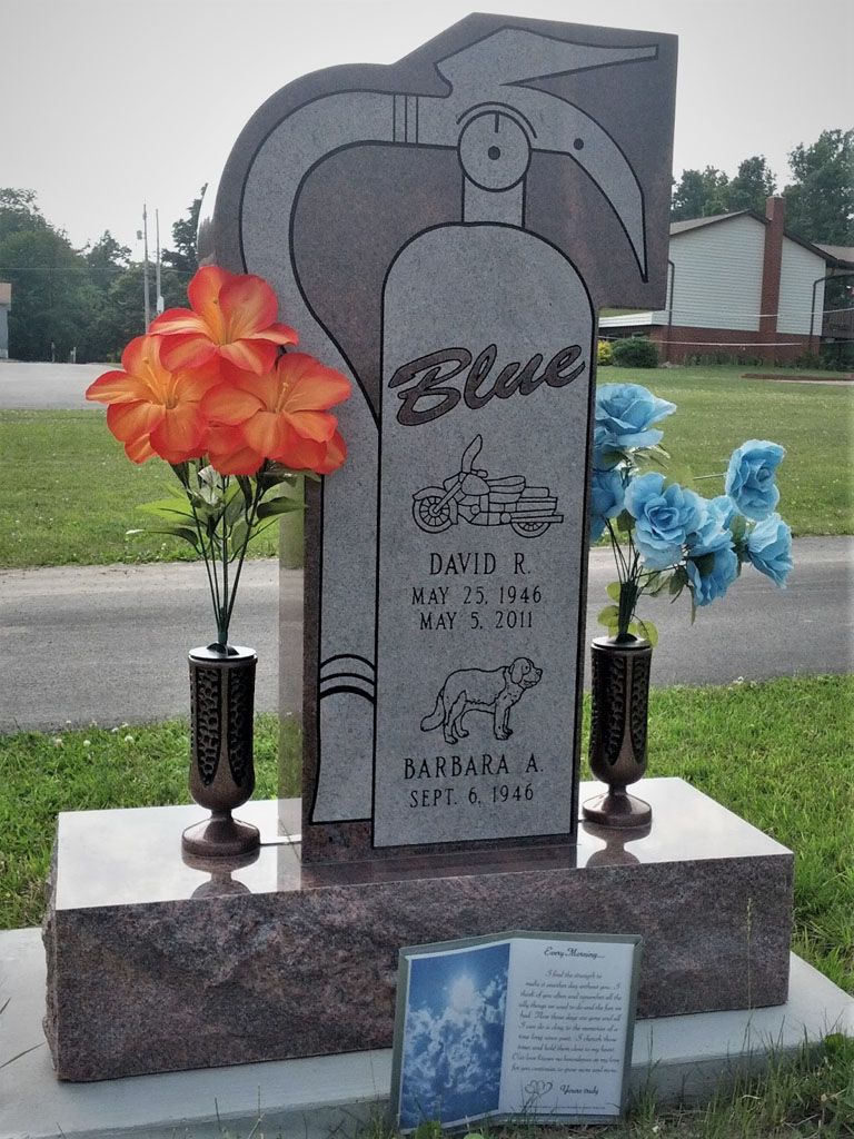 Gravestone with blue flowers and a plaque, set on a stone base in a cemetery.
