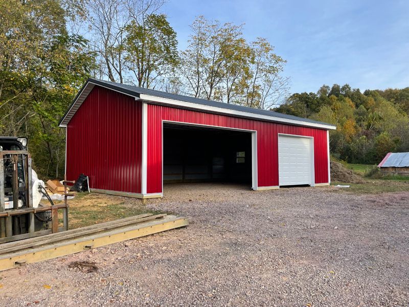 Red barn with large open door, white garage door, gravel driveway, and trees in the background.