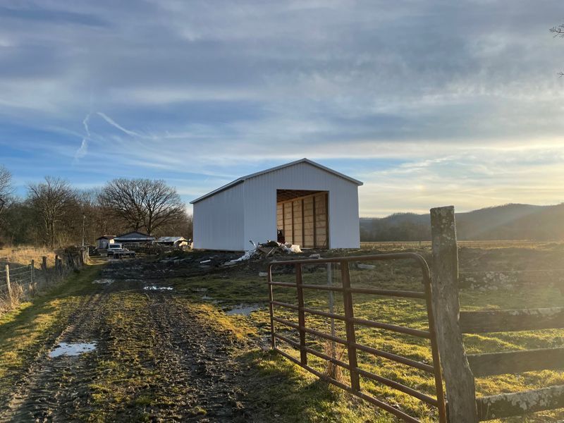 White barn under construction in a muddy field, with a fence in the foreground. Blue sky with clouds.
