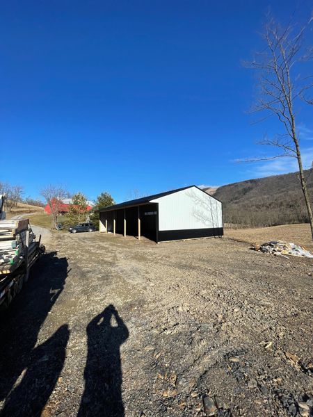 A black-roofed, white-sided barn with open bays sits in a dirt yard under a blue sky.