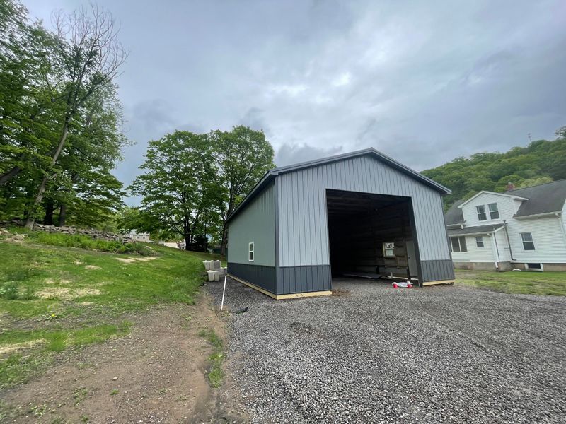 Gray and black metal barn with open front on a gravel driveway, cloudy sky.