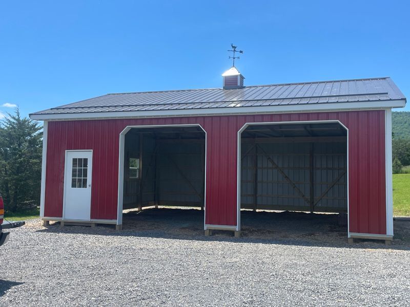 Red and white garage with two bays, a door, and a weather vane on a gravel lot under a blue sky.