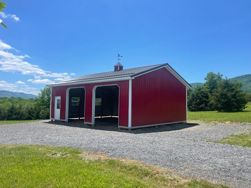 Red barn-style shed with two garage door openings, a white door, and a weathervane, set against a blue sky.