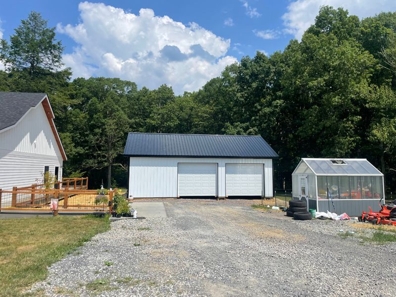 White garage with black roof and two overhead doors, gravel driveway, greenhouse, and trees.