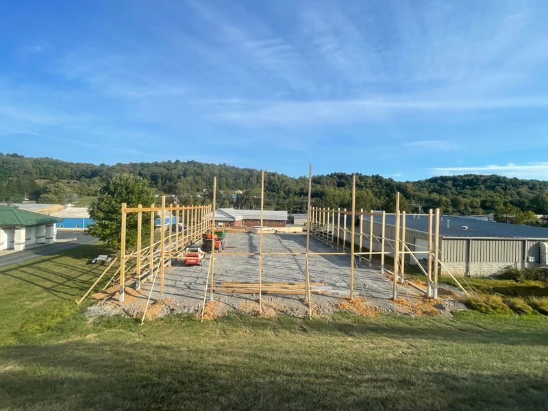 Construction site with wooden frame structure, gravel ground, and surrounding buildings under a blue sky.