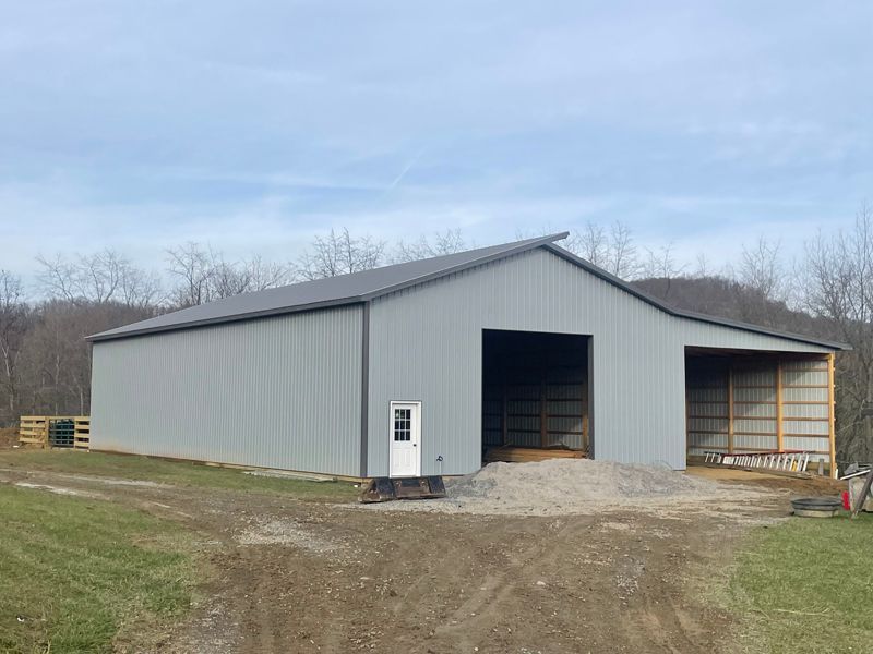 Gray metal barn with open bays, white door, and gravel base on a grassy hillside.