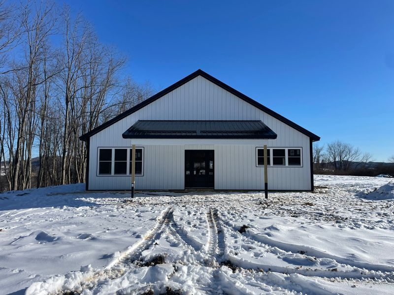 White barn with black trim, windows, and a snowy path leading to the entrance under a clear blue sky.
