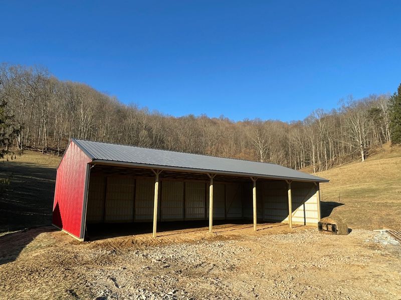 Red and beige open-sided shed with gray roof in a field; trees in background; sunny day.