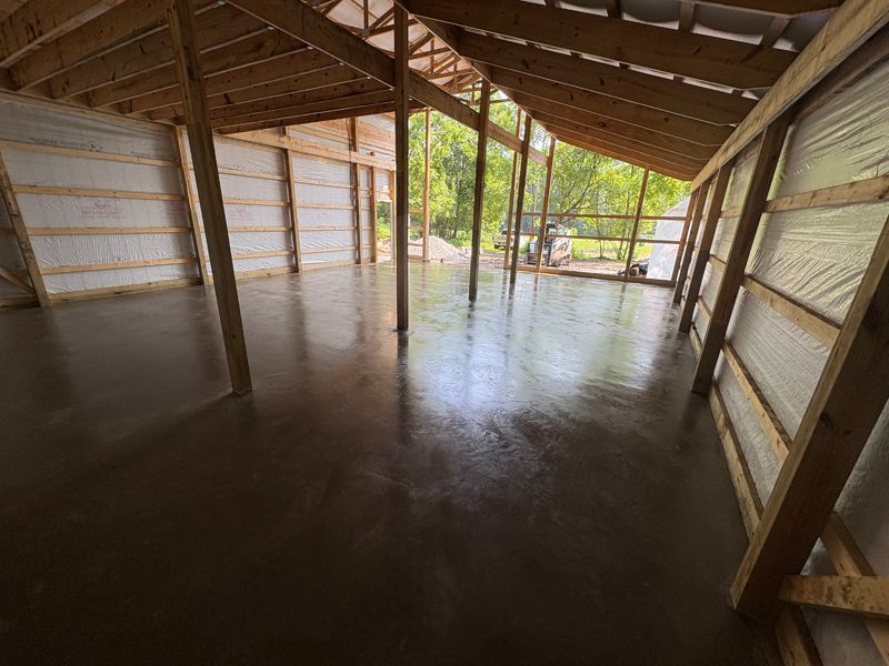 Interior of a building with a fresh, wet concrete floor. Wooden beams frame the space with natural light.