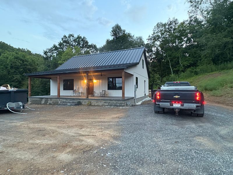 White cabin with dark roof and porch. A truck is parked in front on a gravel driveway.