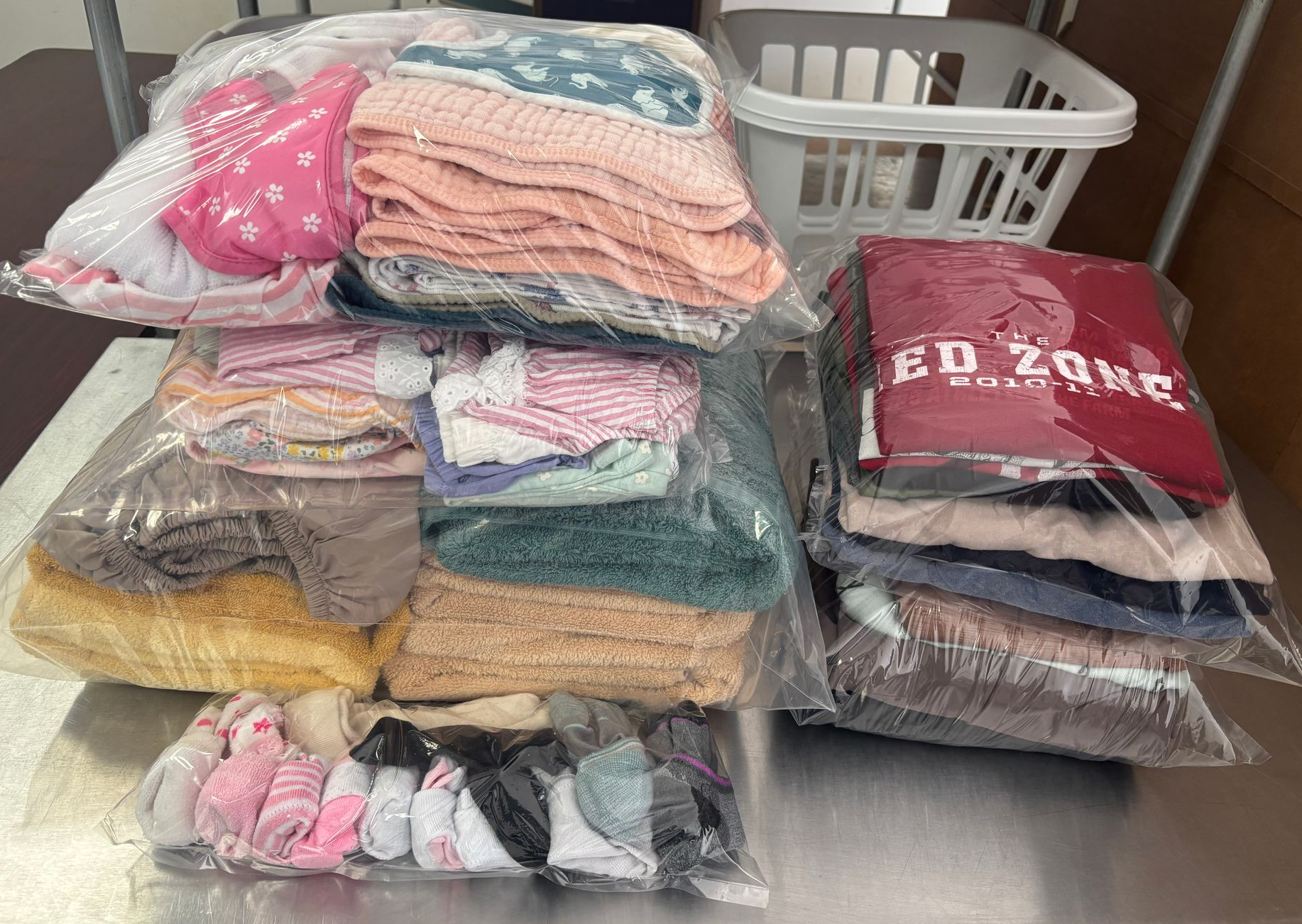 Several clear plastic bags of folded clothing on a metal surface, with a white laundry basket in the background.