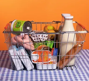 A shopping basket filled with groceries is sitting on a checkered table cloth.