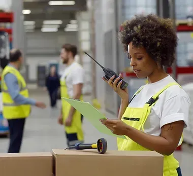 A woman is talking on a walkie talkie in a warehouse while looking at a clipboard.