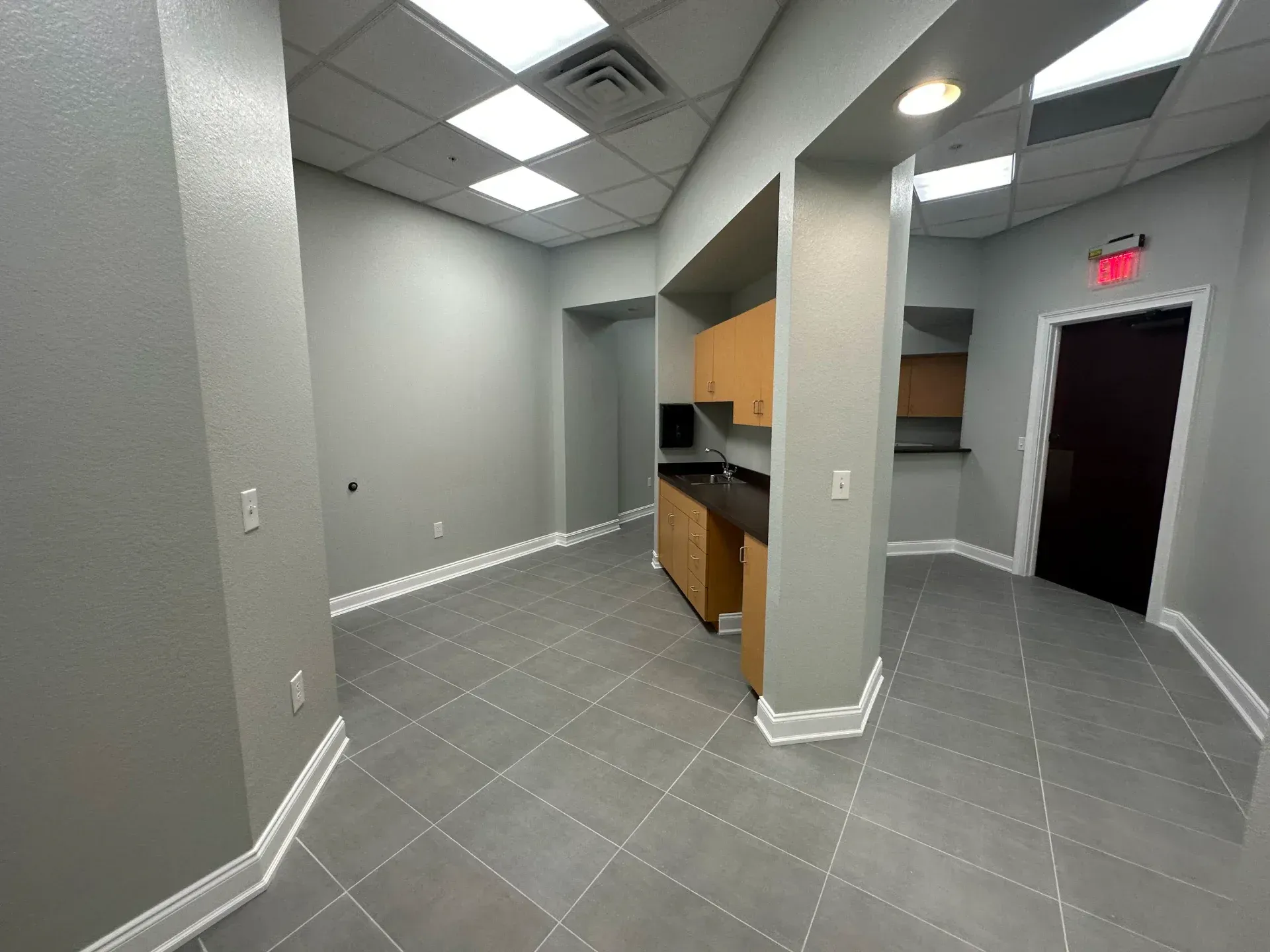 Empty, gray-walled office with cabinets, tiled floor, and ceiling lights; an exit sign is visible.