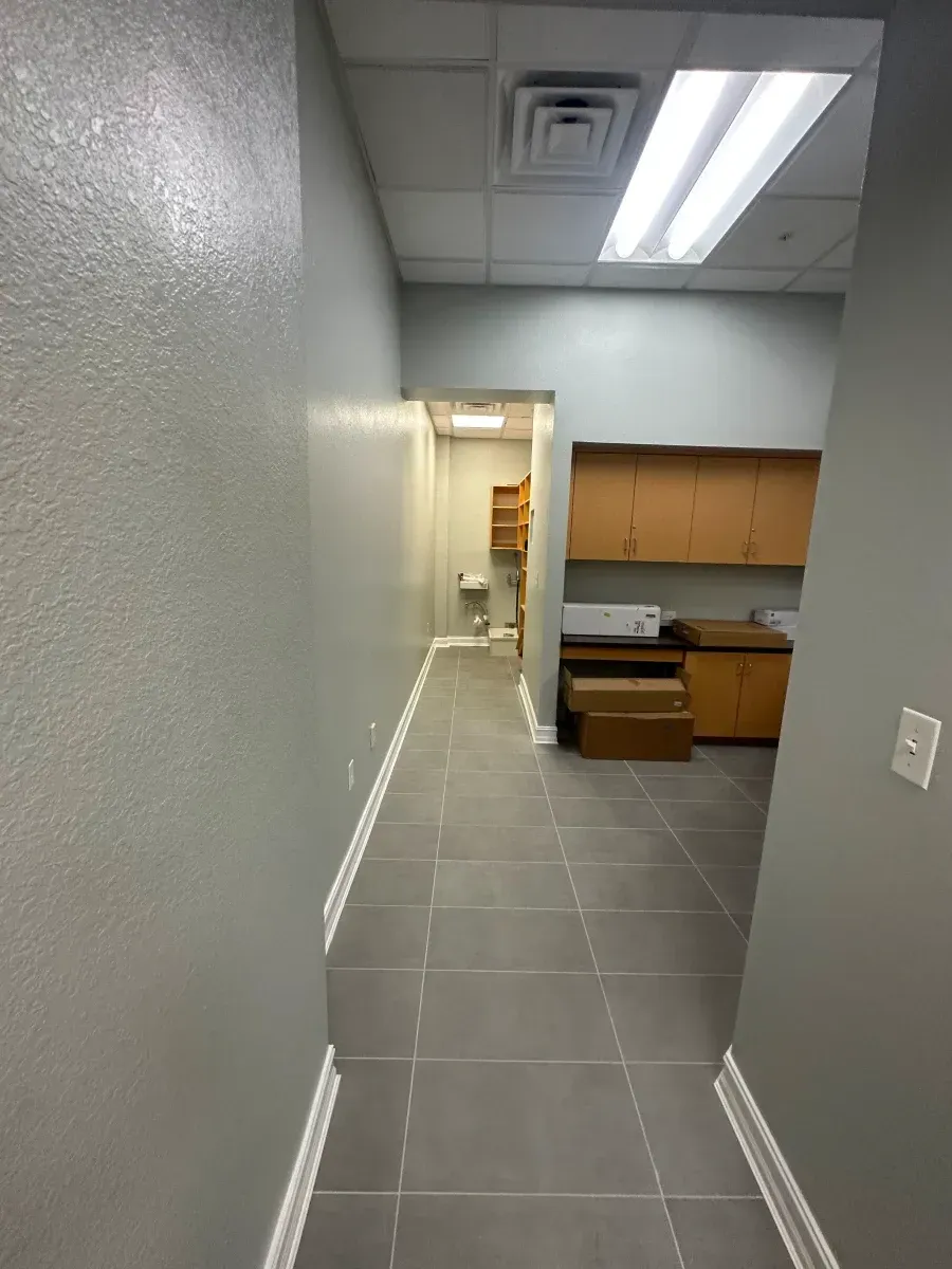 Narrow office hallway with gray walls, tile floor, and a doorway leading to a utility room.