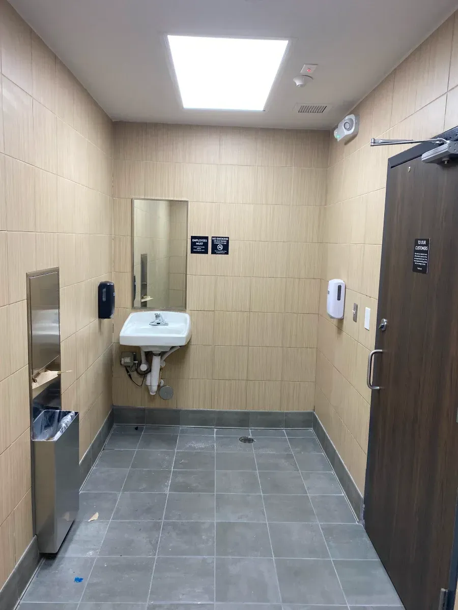 Empty, tiled public restroom. Sink with mirror, soap dispenser, hand dryer, trash can, and closed door.