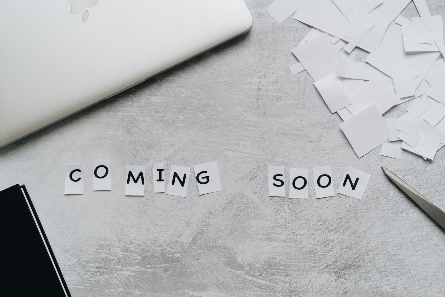 "COMING SOON" spelled out in cut-out letters on a gray surface with laptop and scissors."