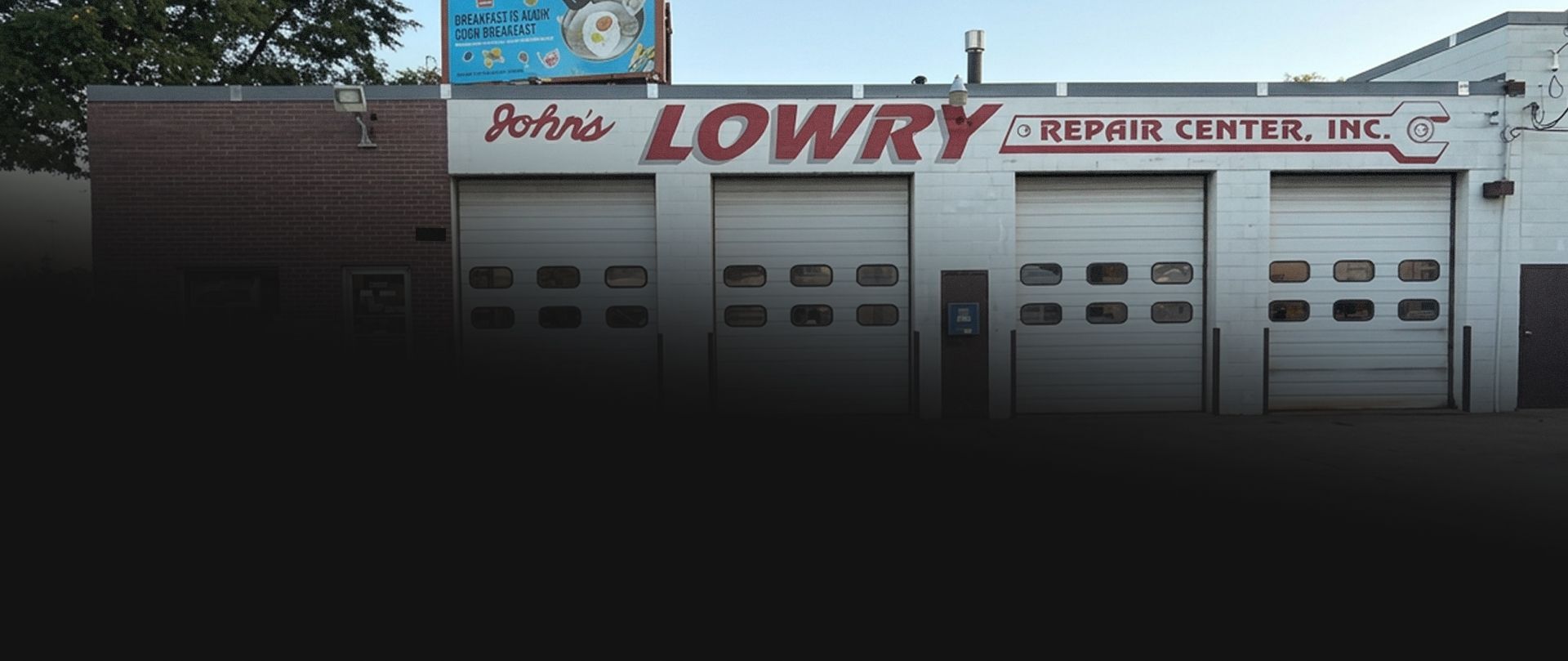 Storefront with red “LOWRY” sign, white garage doors, and a brick wall at dusk | Lowry Repair Center Inc
