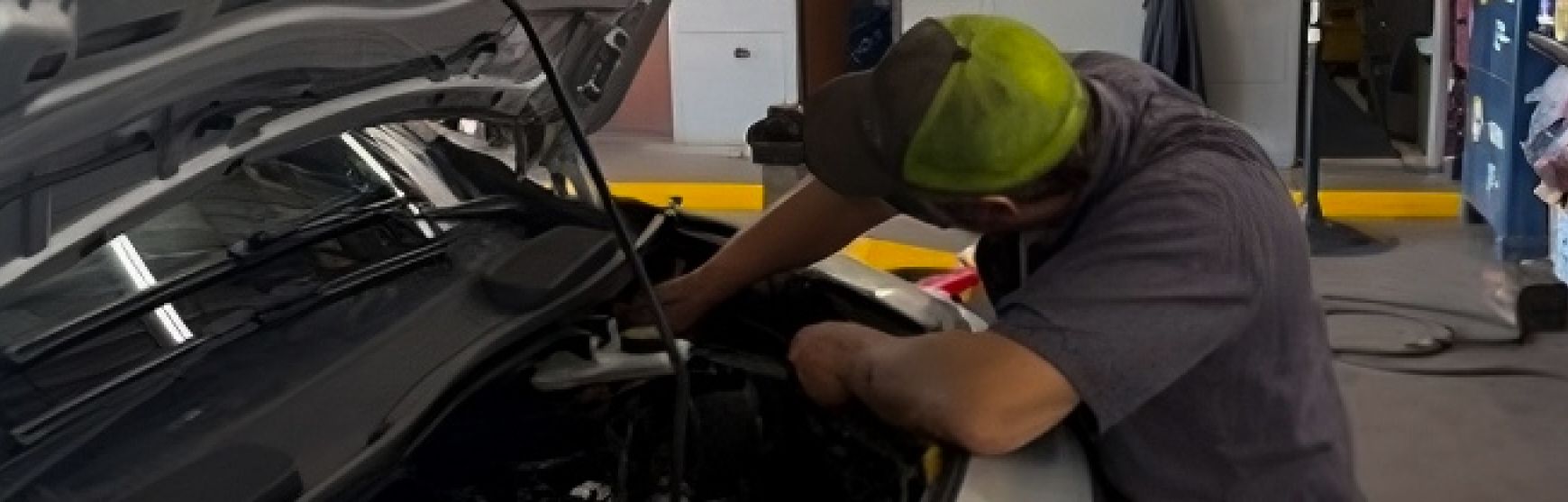 Mechanic working under an open car hood in a garage, wearing a neon cap and gray shirt | Lowry Repair Center Inc

