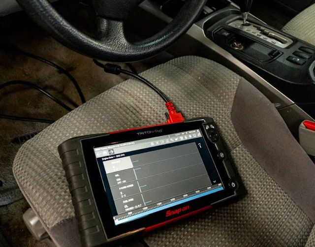 Black tablet with a diagnostic screen sitting on a car seat, with a steering wheel and console nearby | Lowry Repair Center Inc