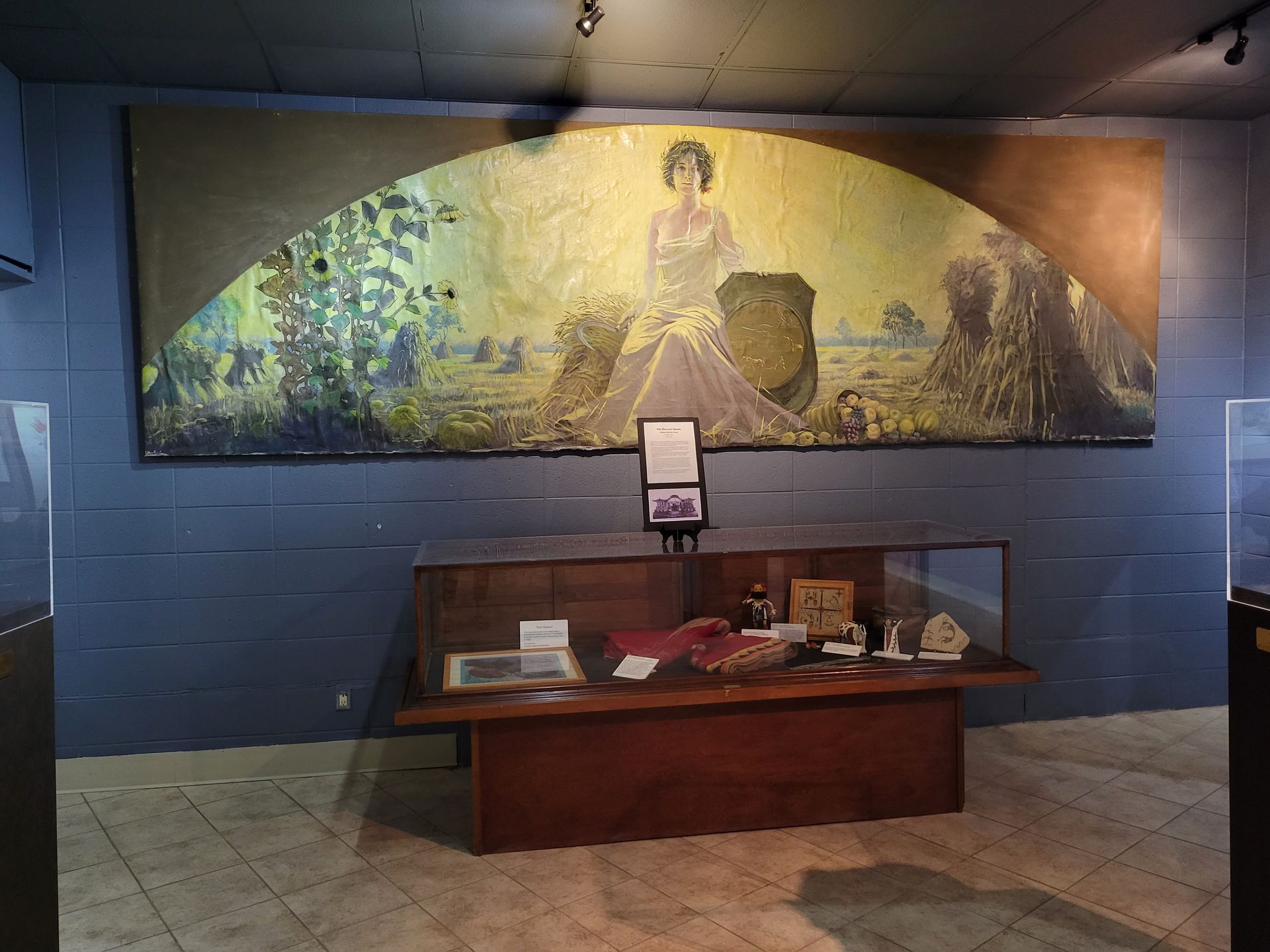A mural of a central figure in a field sits above a display case in a room with blue walls and tiled flooring.