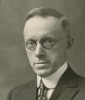 Black-and-white portrait of a man wearing round glasses, a suit, and a tie against a plain background.