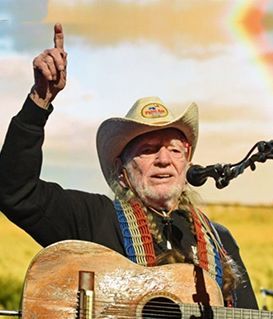 Willie Nelson in a cowboy hat and red-white-blue scarf plays guitar, holding one finger up while performing on stage.
