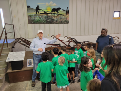 A guide stands before a group in a museum, gesturing toward antique metal plows displayed near a mural of a farmer.