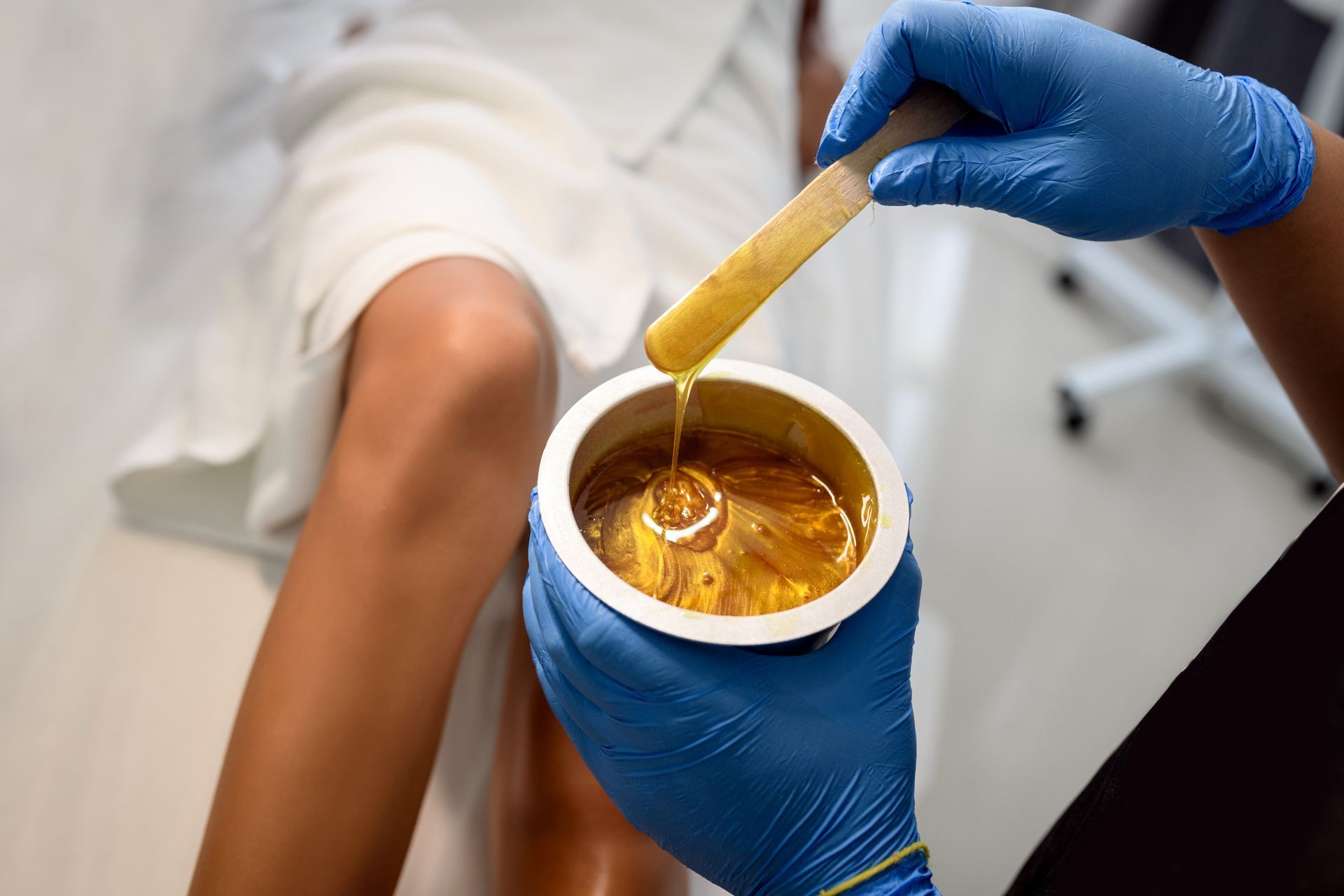 Blue-gloved hands applying wax with a wooden stick to a client’s leg in a spa treatment