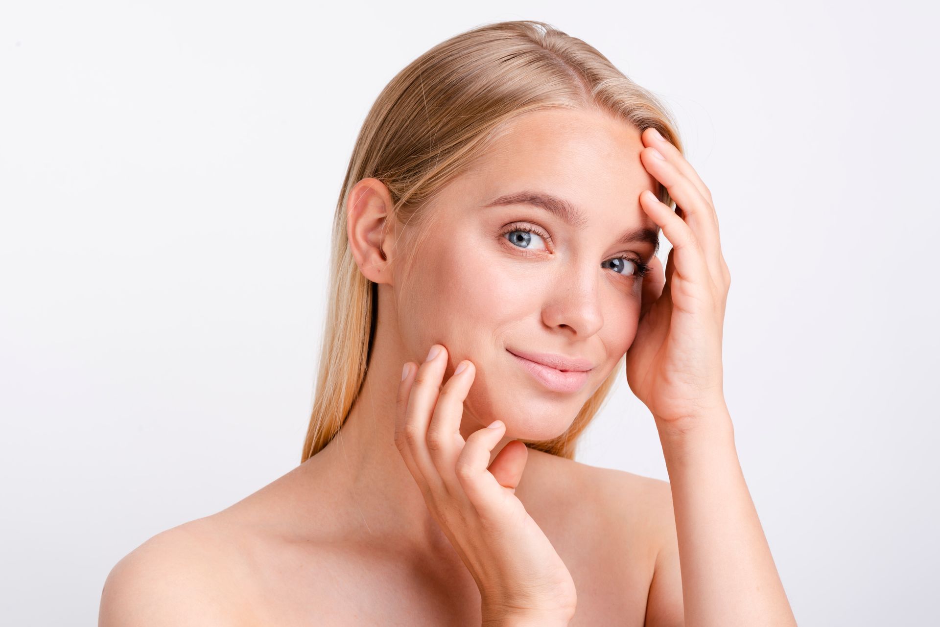 Blonde woman, lightly smiling, touching her face with both hands; white background.