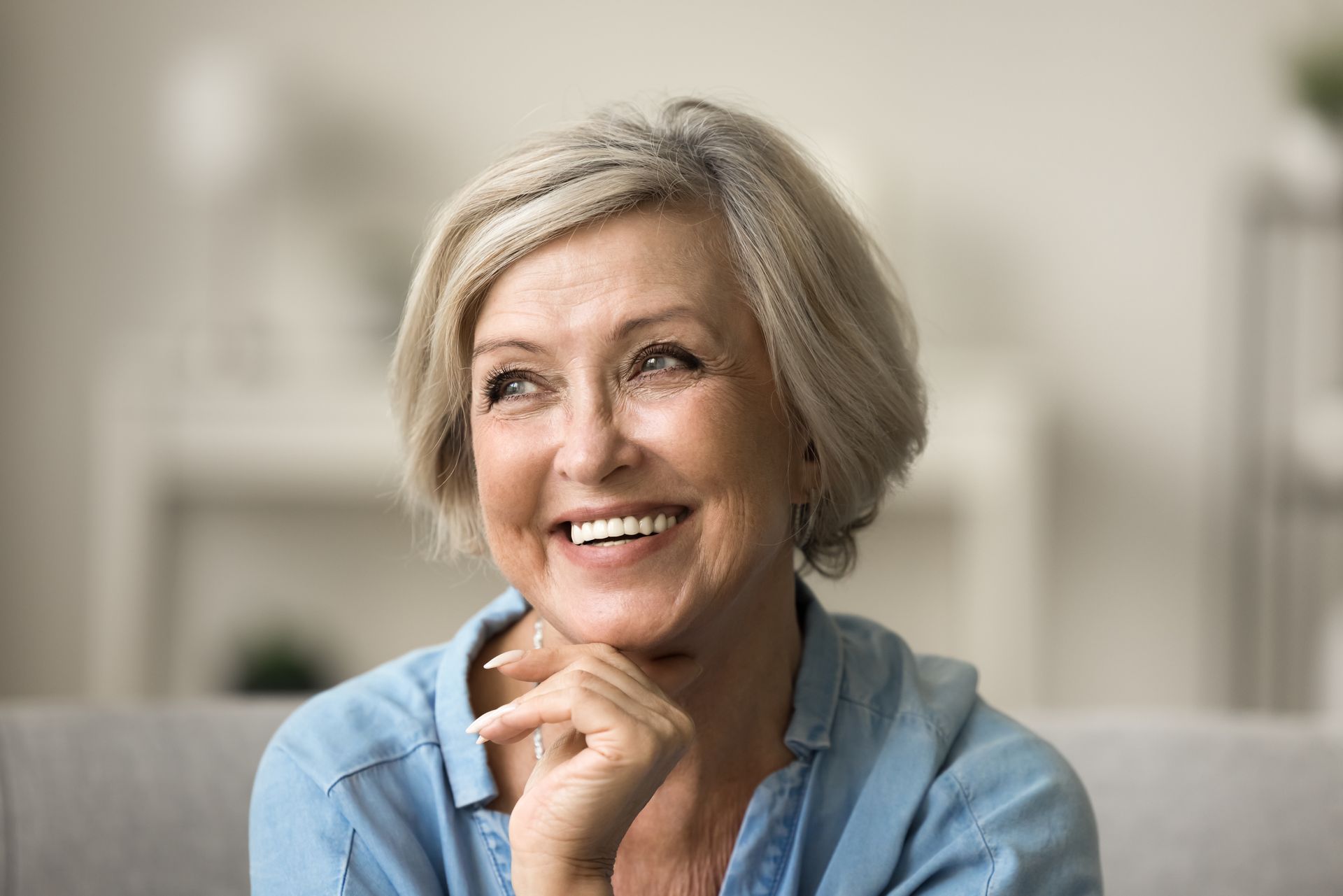 An older woman is smiling while sitting on a couch with her hand on her chin.