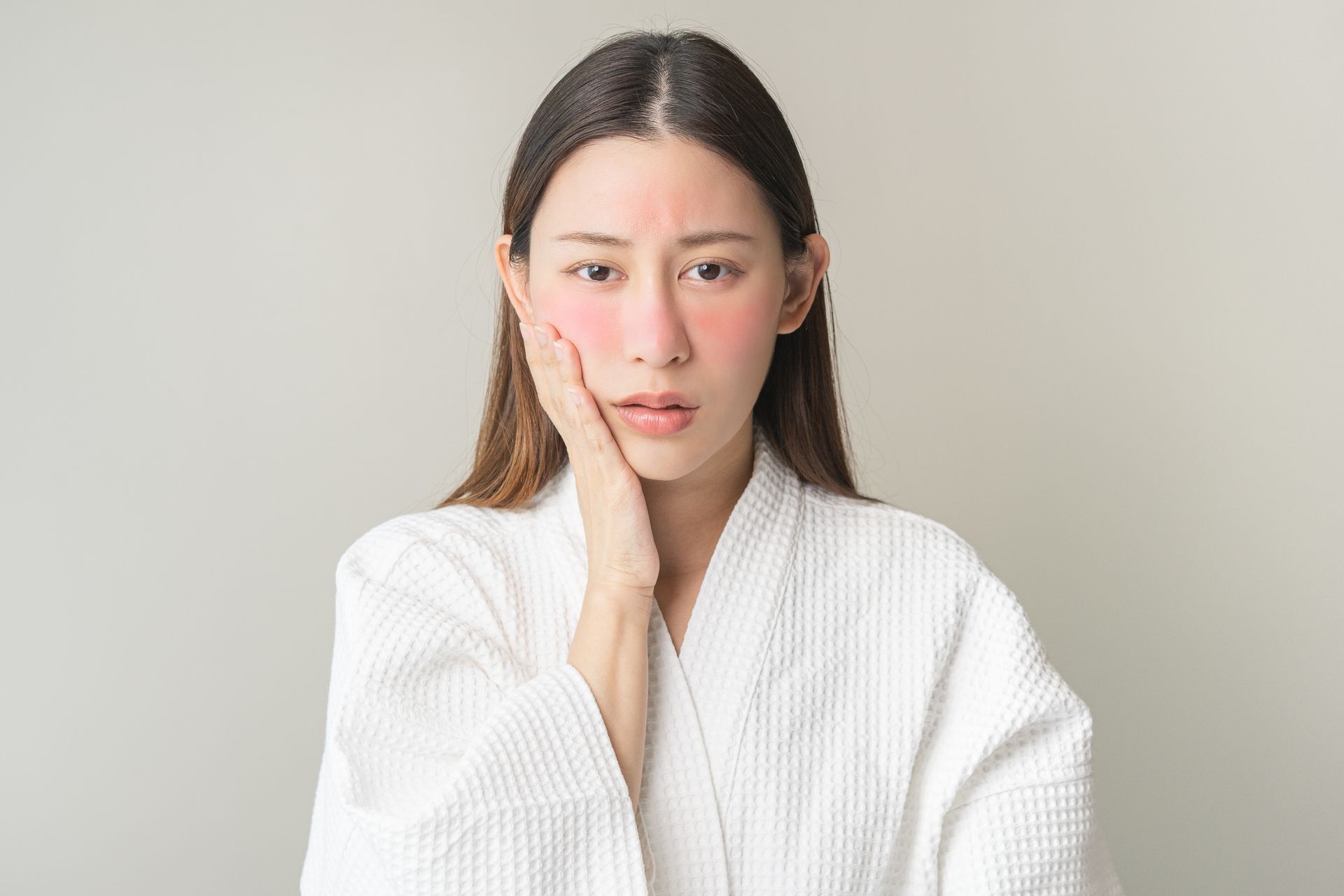 Woman in white robe with red rash on face, touching cheek; neutral background.