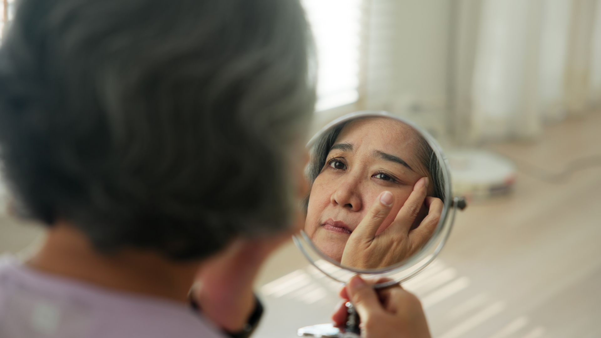 Person looking at their face in a handheld mirror, touching their cheek. Indoors, natural light.