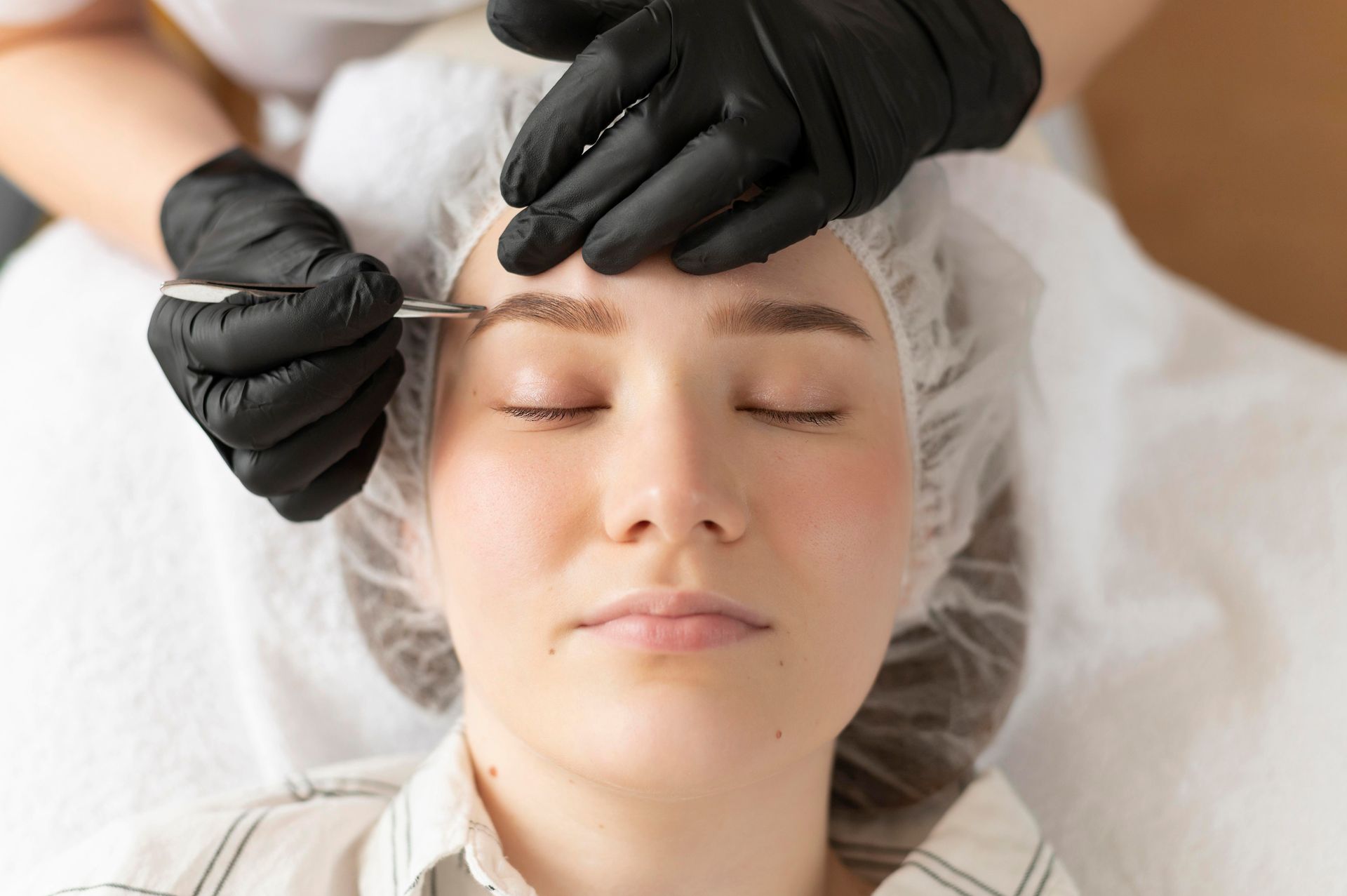 Beautician applying eyebrow treatment to a client lying on a white bed
