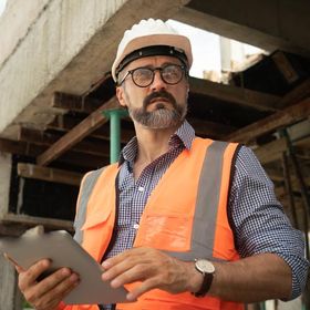Construction worker in hard hat and vest holding a clipboard, inspecting a building site.