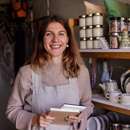 Woman smiling, holding notepad, standing in a shop with shelves of candles and ceramics.