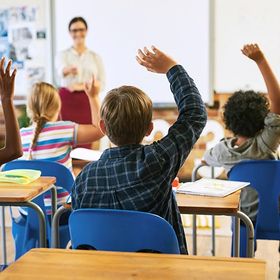 Students in a classroom raise hands to answer a question, teacher in background.