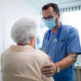 Doctor in scrubs with stethoscope comforts a patient, touching their shoulder. They are in a medical room.