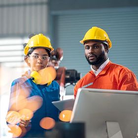 Two workers wearing hard hats and work clothing in a factory setting.