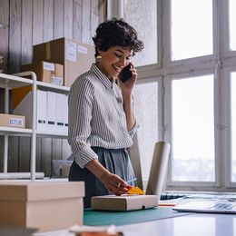 Woman in striped shirt on phone, packing order in a home office with boxes and a window.