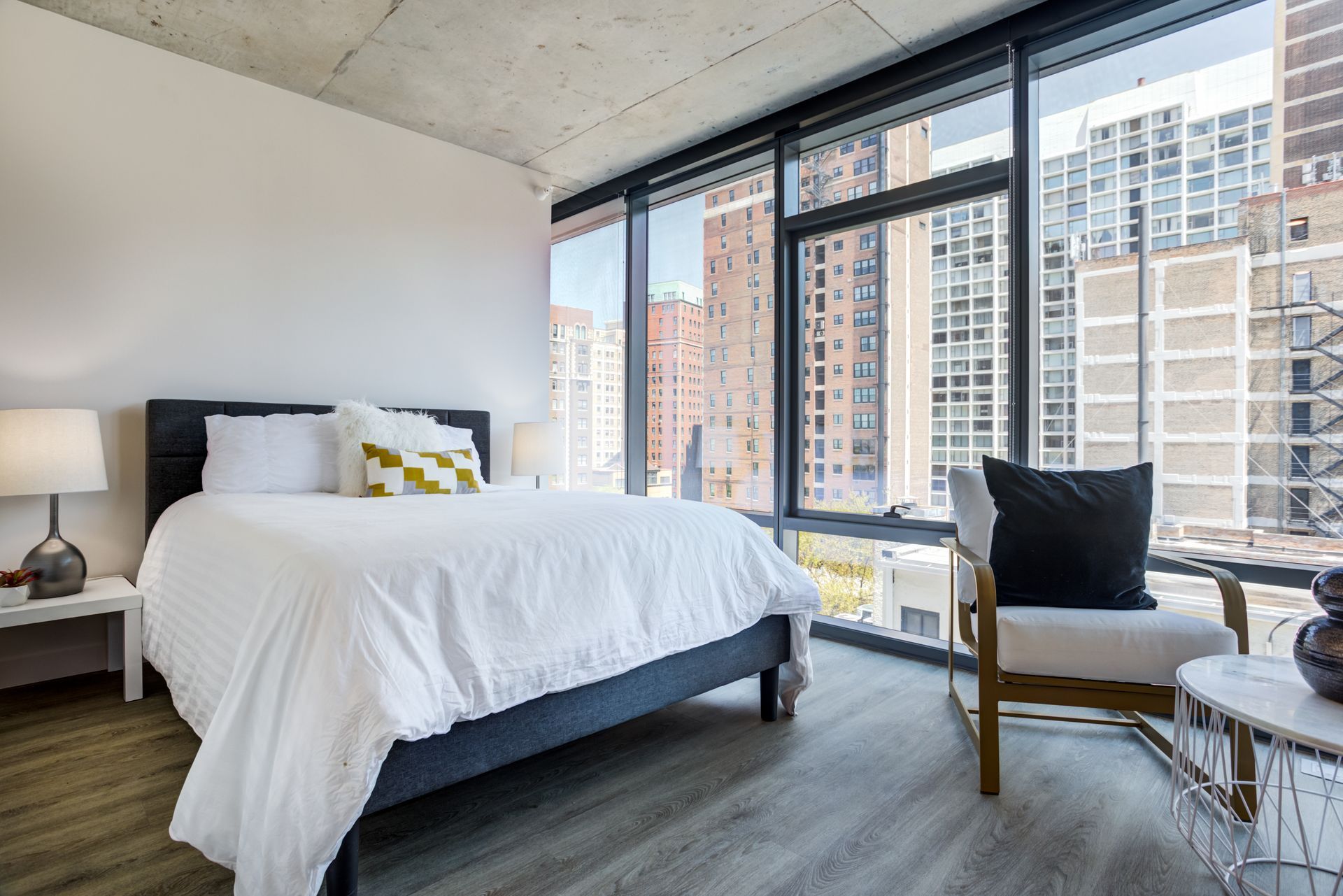 A bedroom with a king size bed , chair , lamps and large windows.