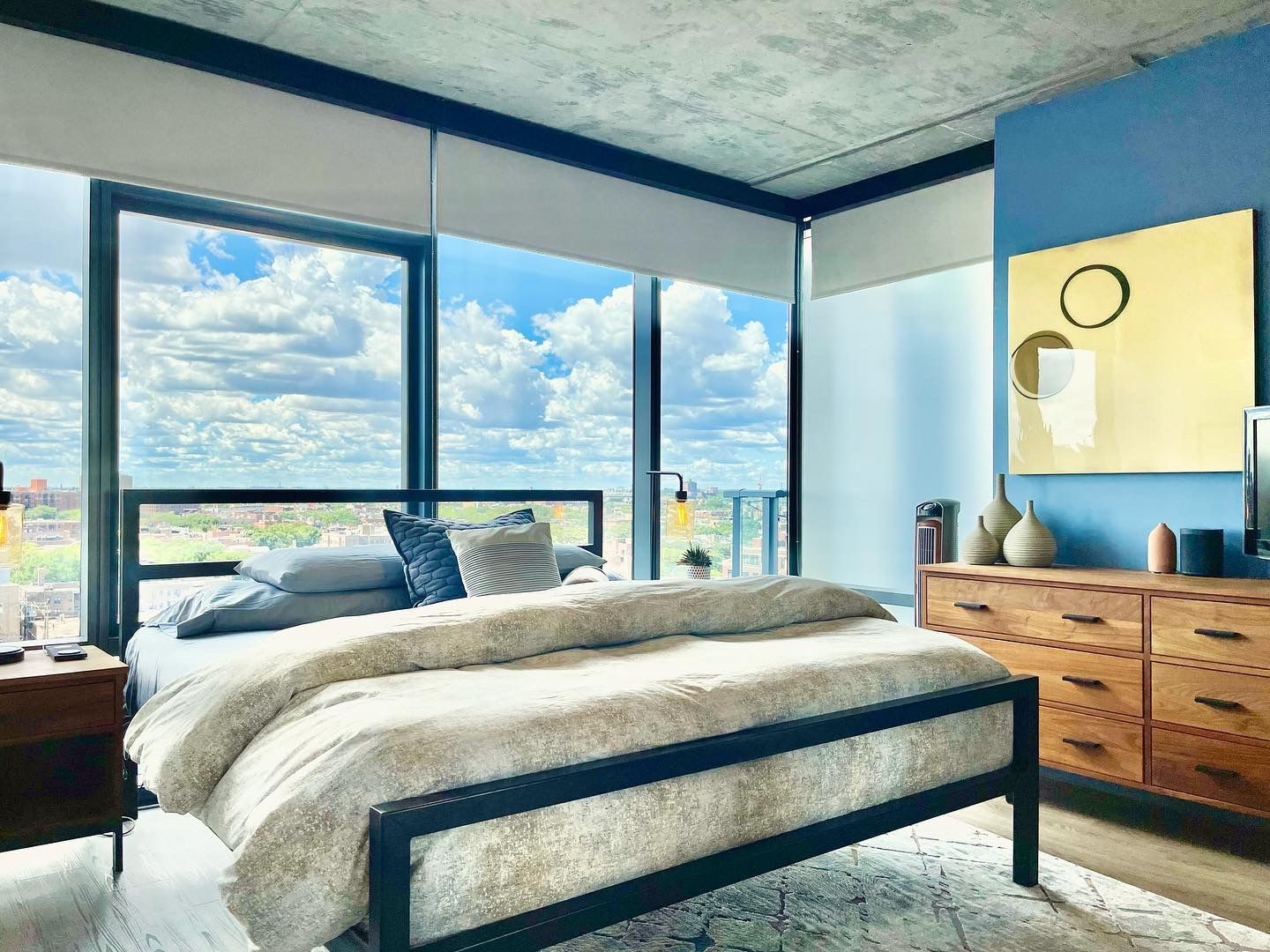 A bedroom with a bed , dresser , nightstand and large windows.