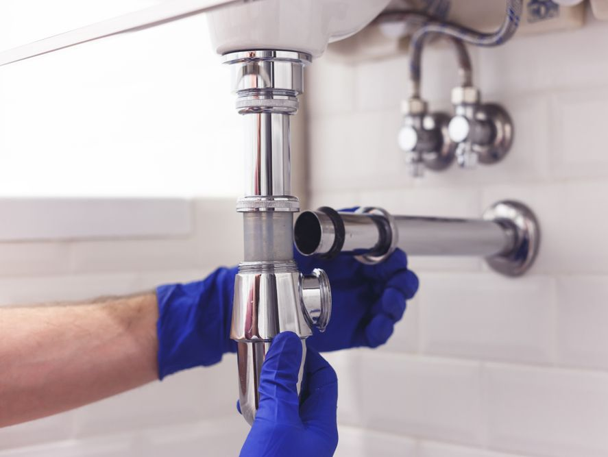Plumber in Blue Gloves Attaching Pipes Under a White Sink in a Tiled Bathroom — Full On Plumbing & Gas in Nightcliff, NT
