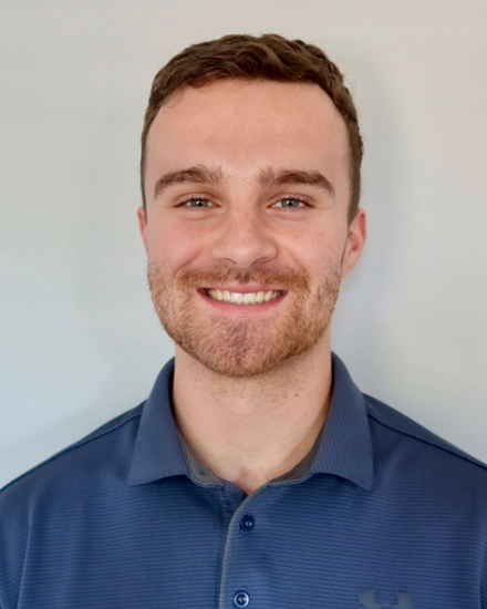 Man with brown hair and stubble smiles, wearing a blue polo shirt, against a gray wall.