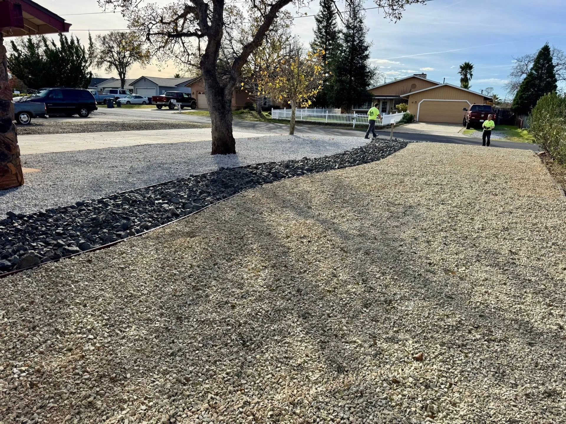 Gravel driveway with two workers. Light and dark gravel areas separated by a line. Houses and trees in the background.